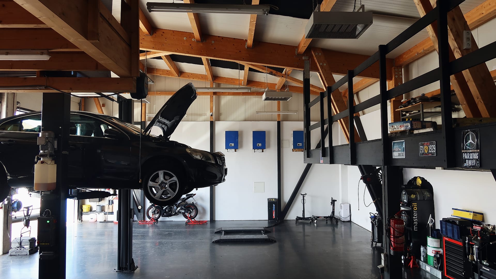 Werkstatthalle am Tag: ein dunkles Auto mit offener Motorhaube auf der Hebebühne, rechts Werkzeugwagen und Ölfässer unter einer Empore