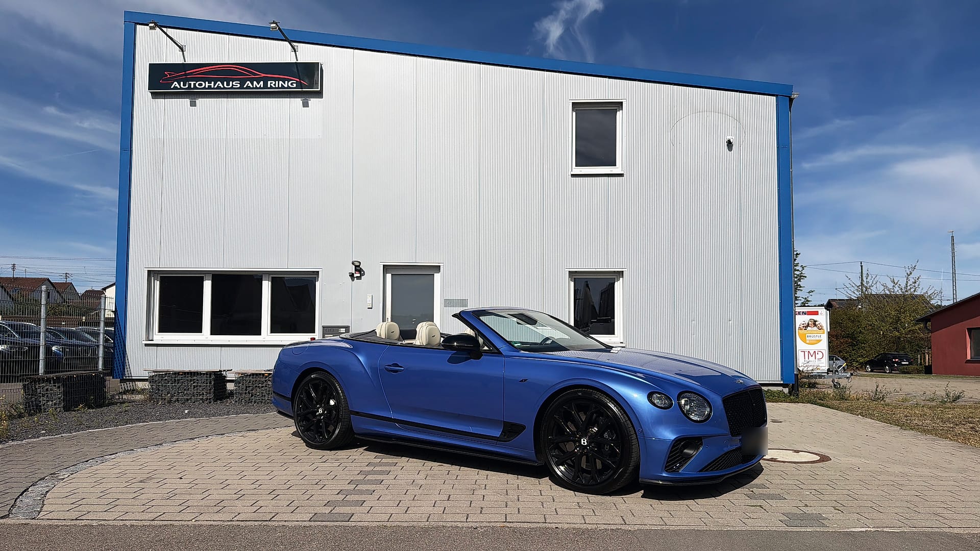 Ein blaues Bentley Continental GTC Cabrio vor der Halle des Autohauses am Ring, darüber das Schild an der Fassade