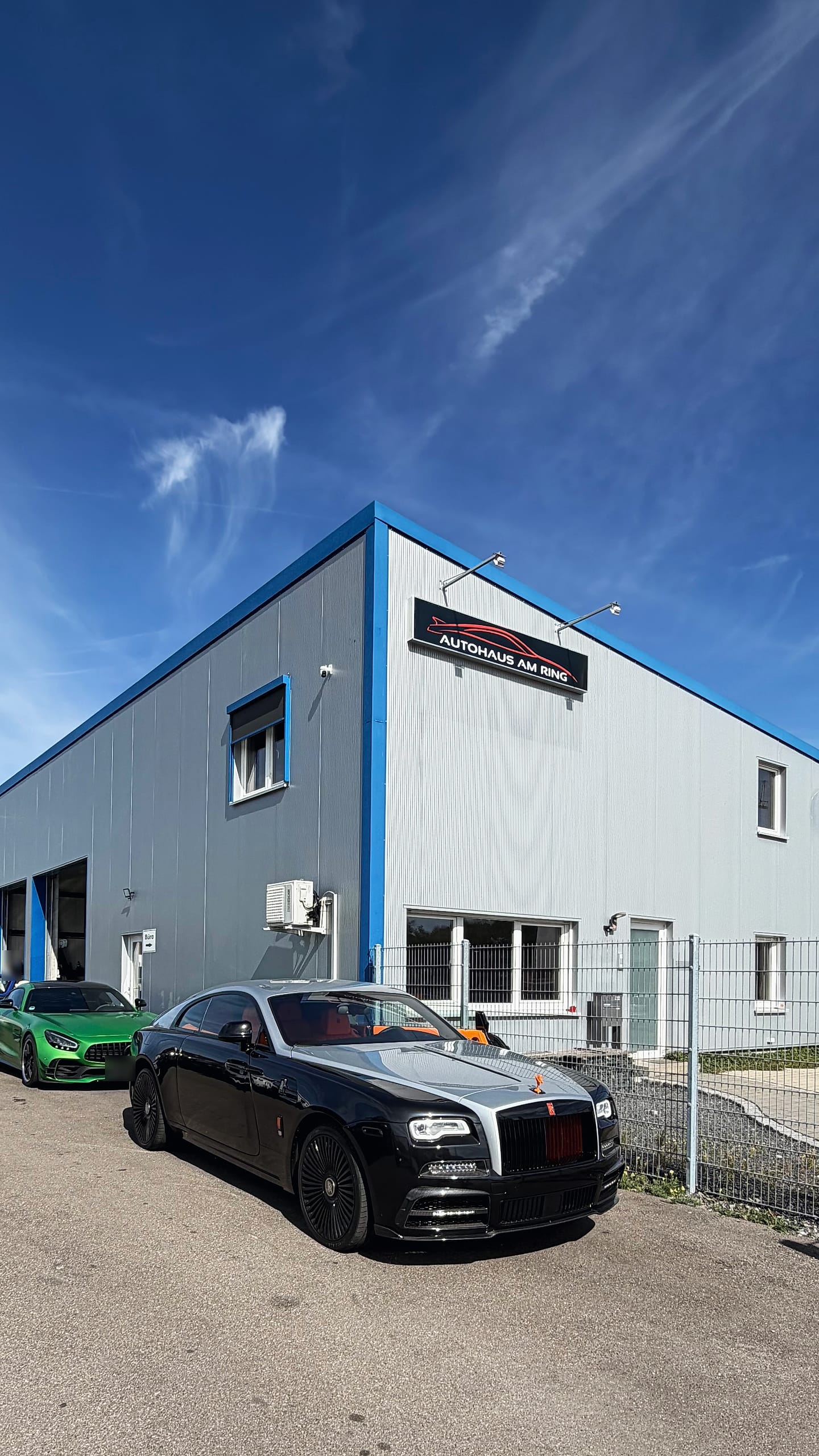 Die Halle über Eck unter blauem Himmel, davor ein Rolls-Royce und ein grüner Mercedes-AMG