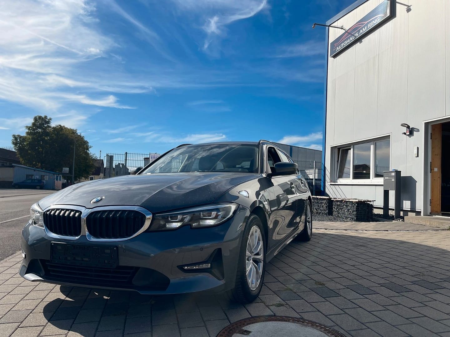 Grauer BMW 3er Touring auf dem Pflaster vor der Halle, an der Fassade das Schild „Autohaus am Ring“